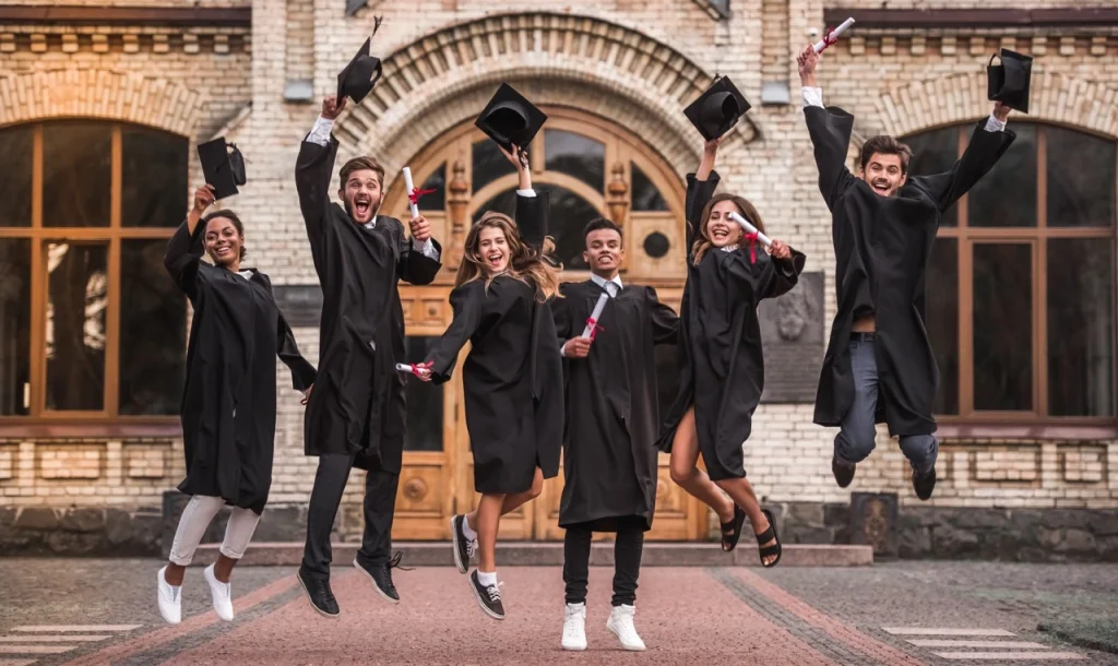 Group of happy graduates in black academic gowns jumping in excitement while holding their diplomas and caps in front of a historic building.