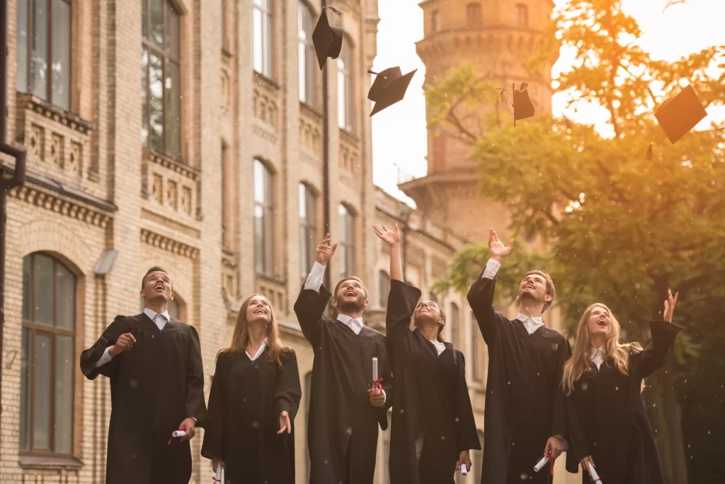 Group Of Graduates In Academic Gowns Joyfully Throwing Their Caps Into The Air Outside A Historic Building During Their Graduation Celebration.